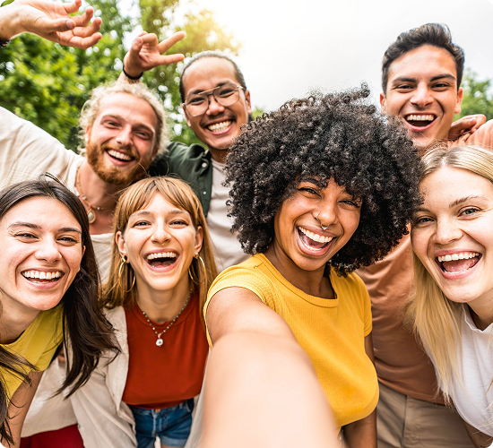 A group of seven diverse people smiling and taking a selfie outdoors, with greenery in the background.