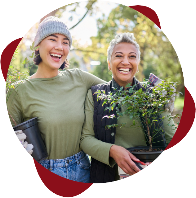 Two people smiling outdoors, holding potted plants. One wears a gray beanie and green shirt, the other a black vest and green shirt. Trees and sunlight are in the background.
