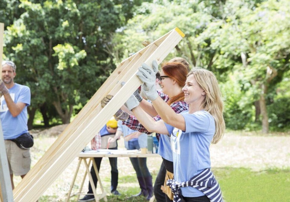 People in casual work attire assemble wooden frames outdoors, surrounded by trees.
