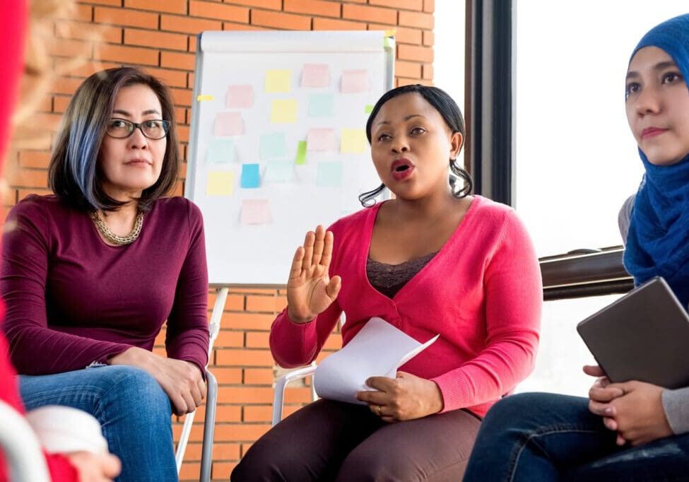 Three women sit in a discussion circle. One woman speaks while holding papers. A flipchart with sticky notes is in the background.